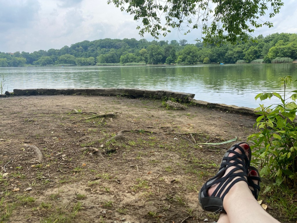 A small lake surrounded by trees sits just beyond a pair of feet in gladiator sandals crossed relaxed at the ankle. 