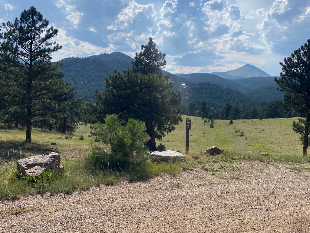 A gravel parking lot sits next to a green field in front of a set of pine tree covered mountains. The sky is sunny with white fluffy clouds.