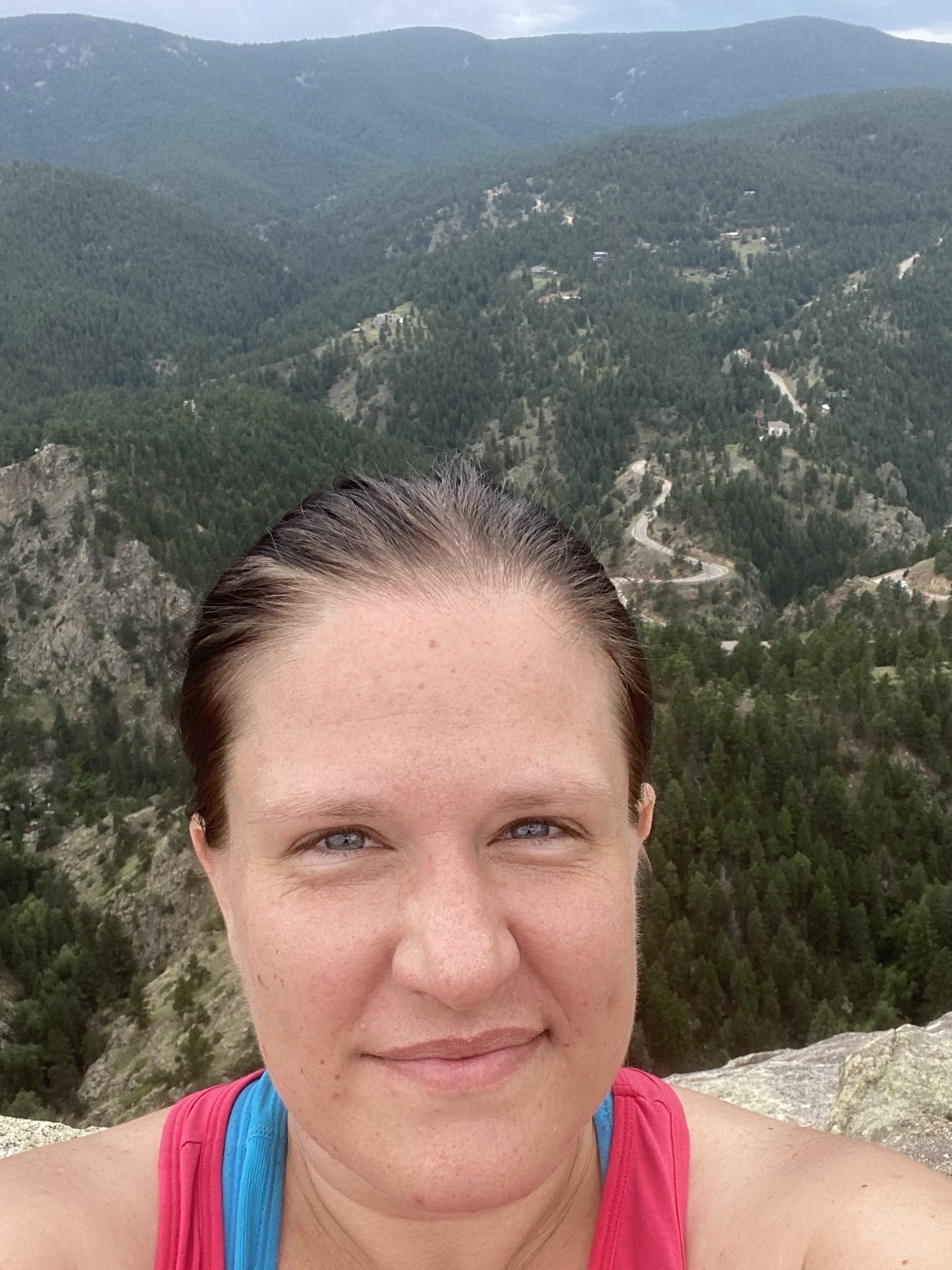 Selfie of a makeup free mid-30's woman on a rock above a beautiful view of the pine tree covered valley below. She's kinda sweaty, but don't mistake that for a tough hike. She just gets tired quickly.